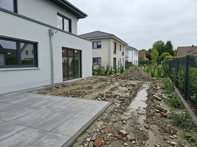 Rohe Baustellenfläche hinter einem Neubau mit Fahrspuren, Pfützen und Bauschutt
